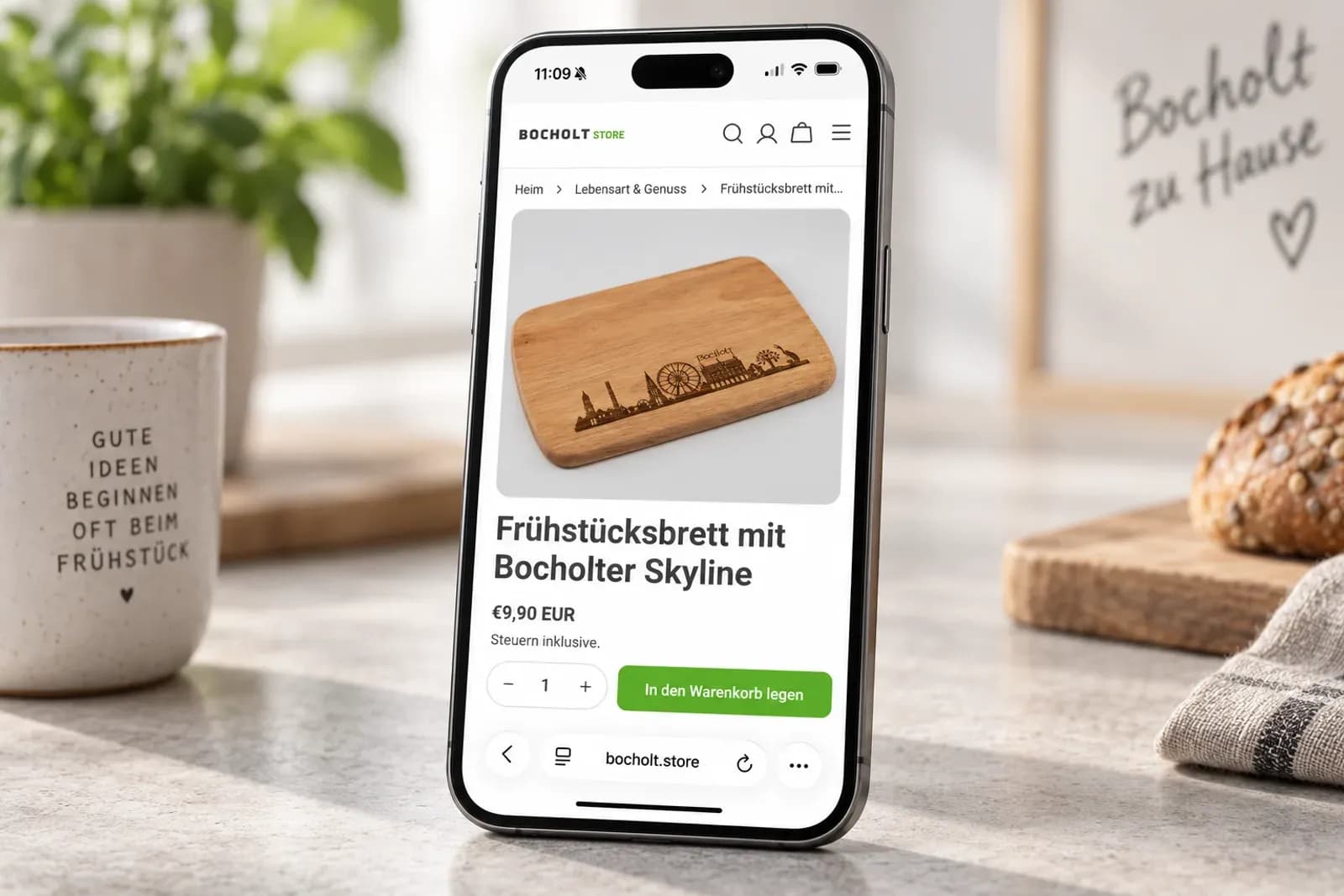 Smartphone mit der Produktseite eines Frühstücksbretts mit Bocholter Skyline auf bocholt.store, daneben Tasse und Brot