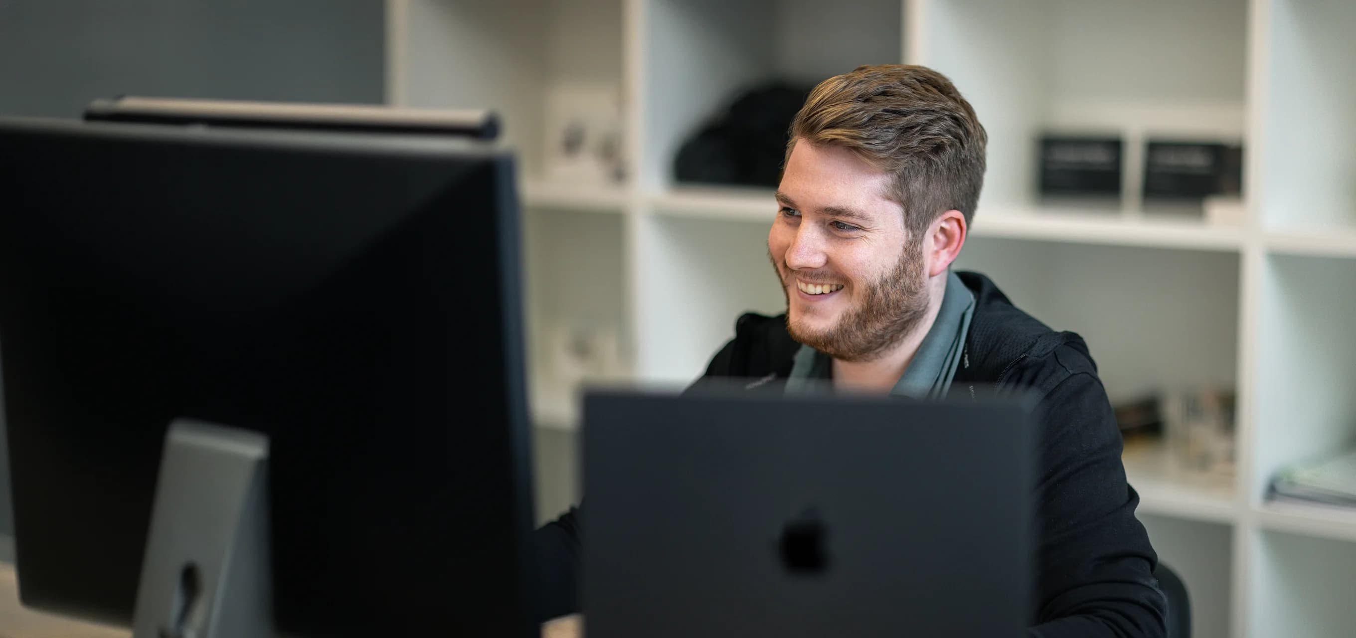 Ein Kollege von skeon digital lacht am Arbeitsplatz zwischen Monitor und Laptop