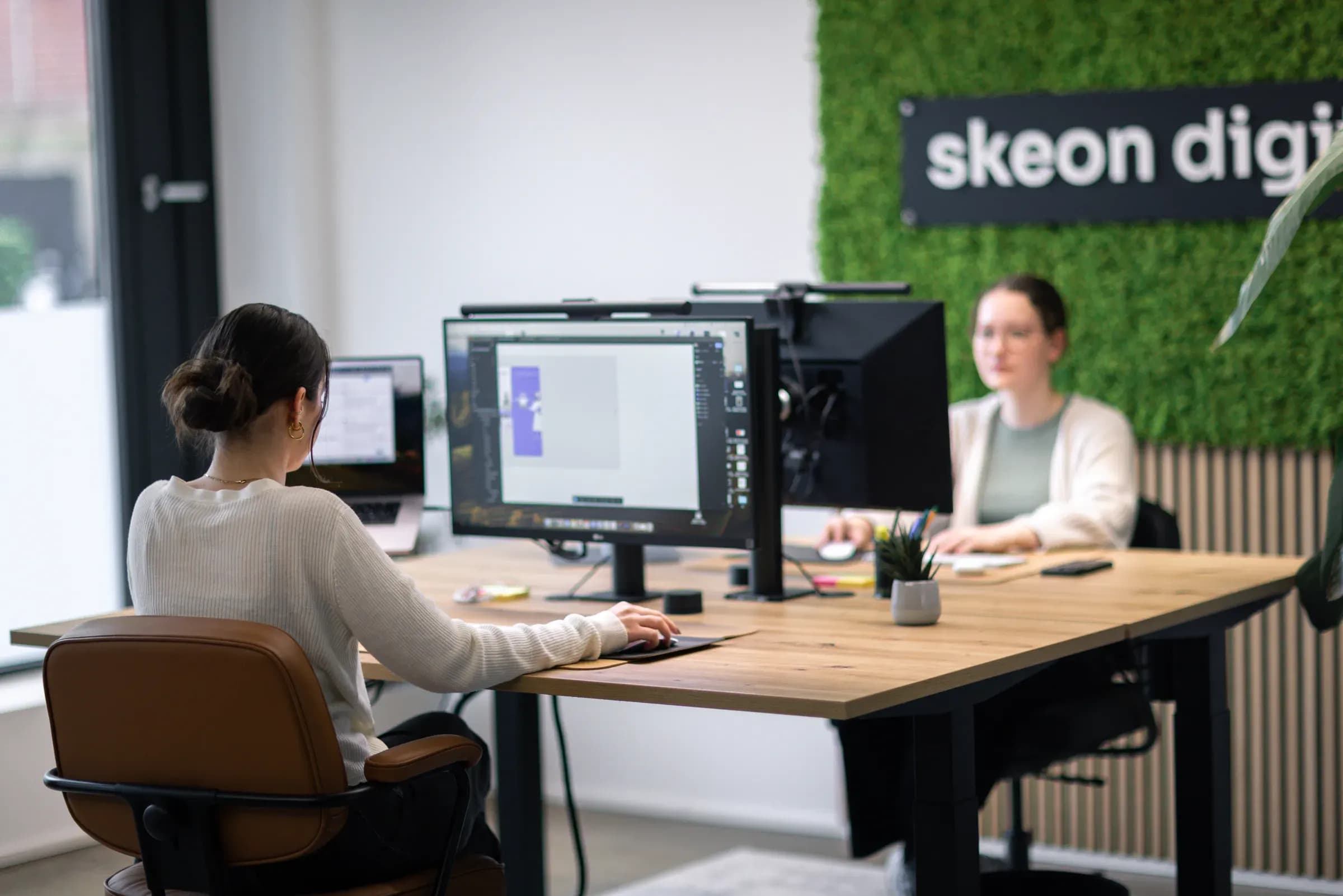 Zwei Kolleginnen an Monitoren im Büro von skeon digital, im Hintergrund die Mooswand mit Logo