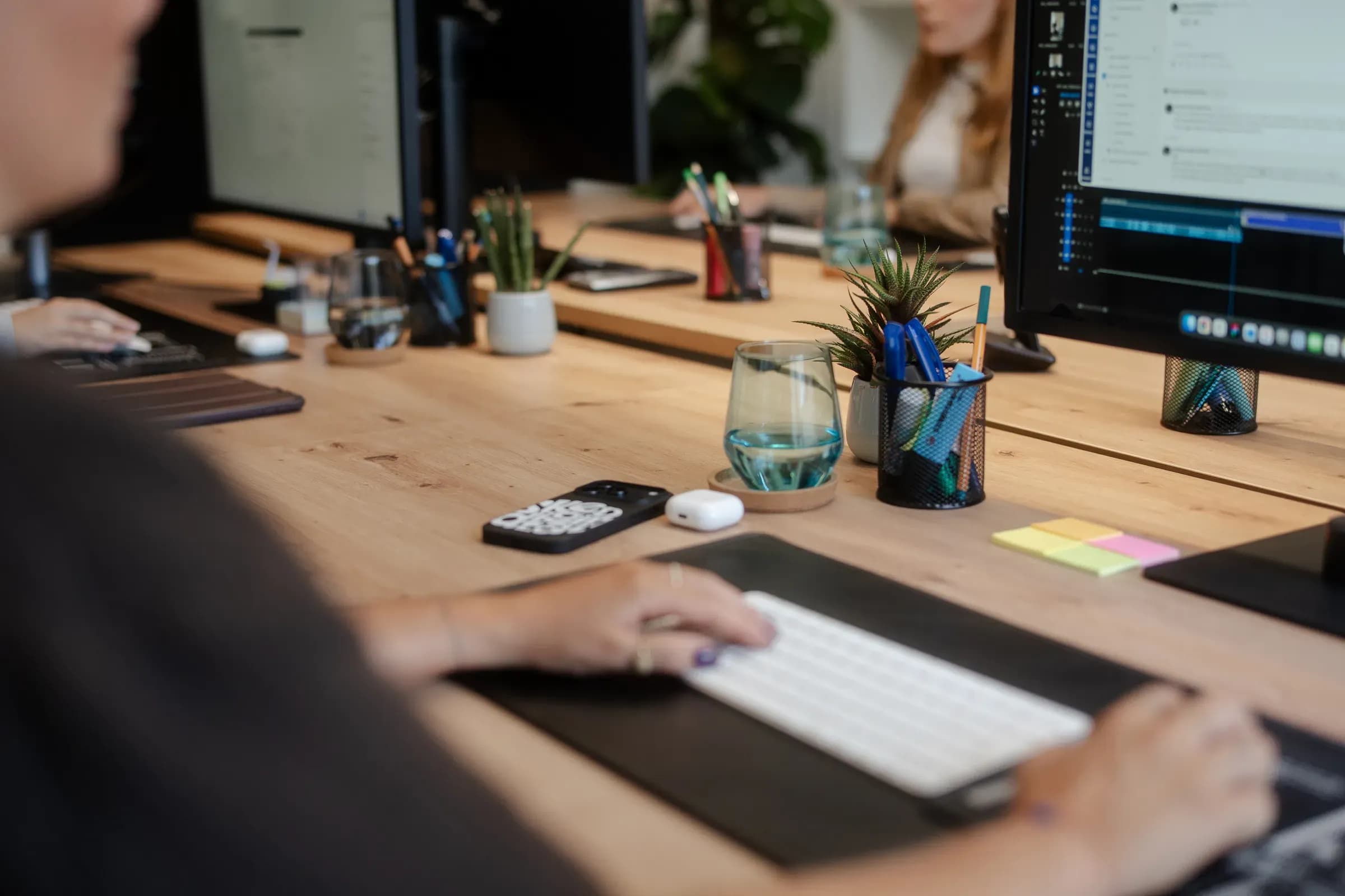 Hände auf der Tastatur am Schreibtisch, im Hintergrund Monitore mit Texten