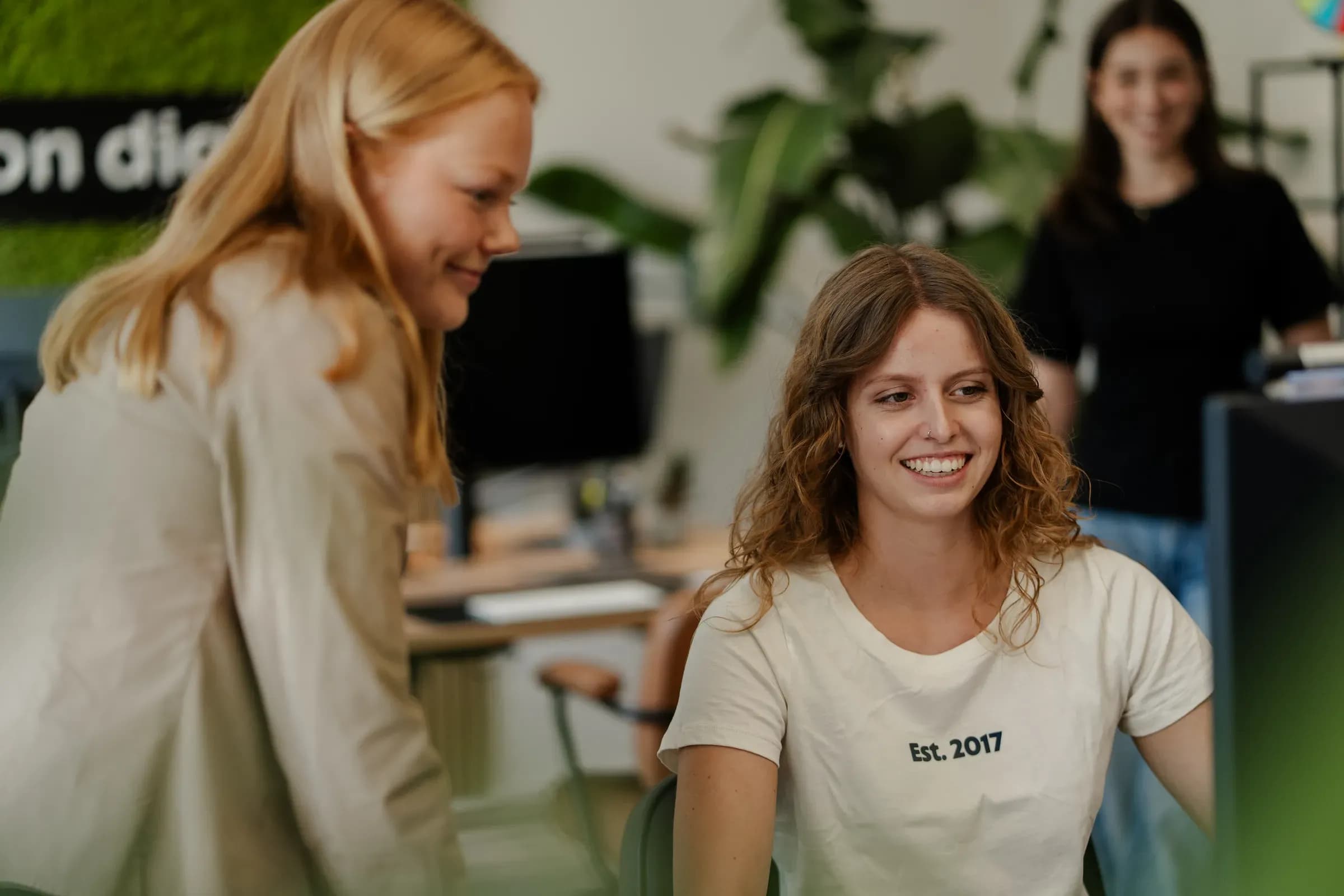Zwei Kolleginnen von skeon digital im Gespräch am Schreibtisch, eine lächelt in die Kamera