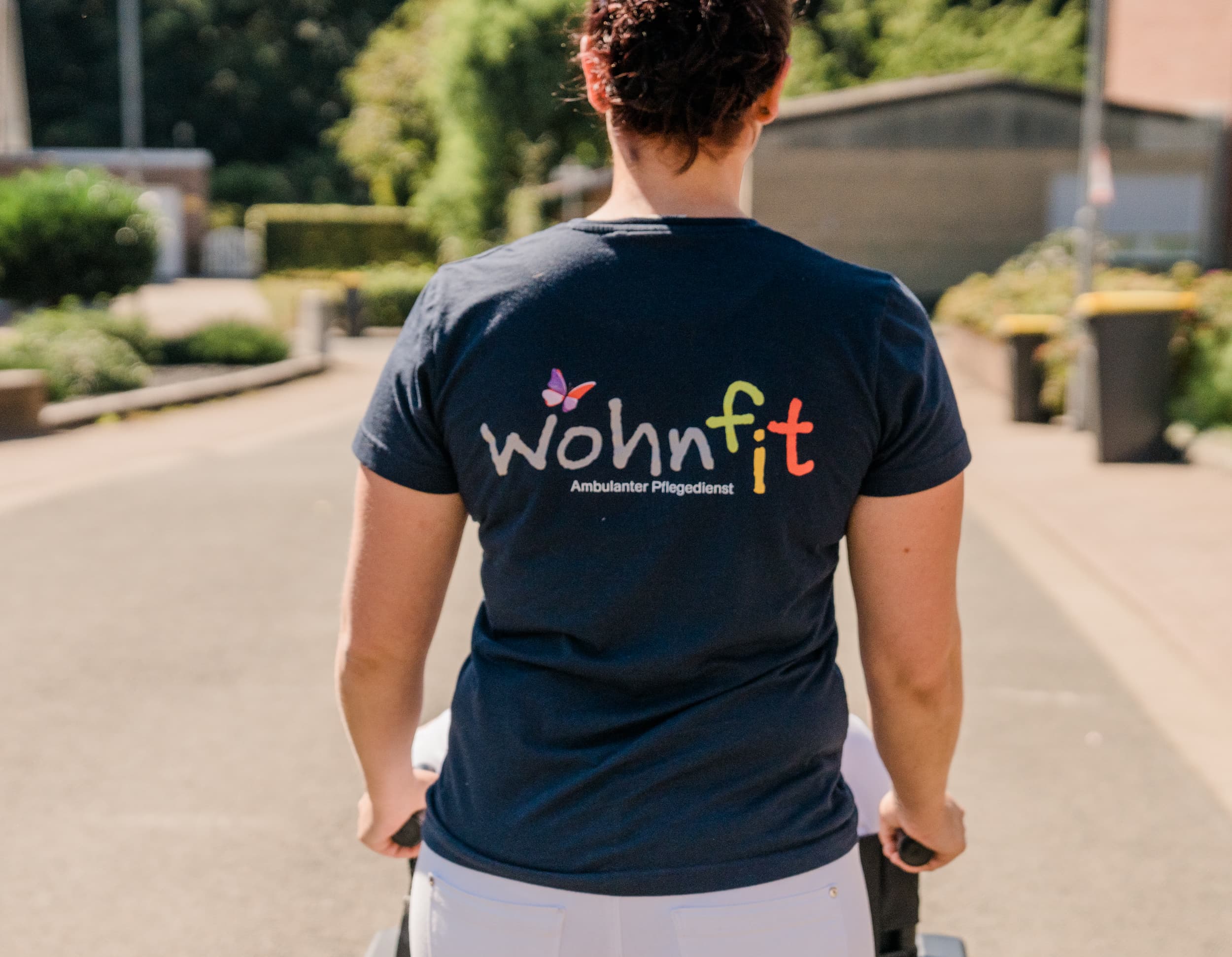 Pflegekraft im T-Shirt mit wohnfit-Logo, von hinten, schiebt einen Rollstuhl.