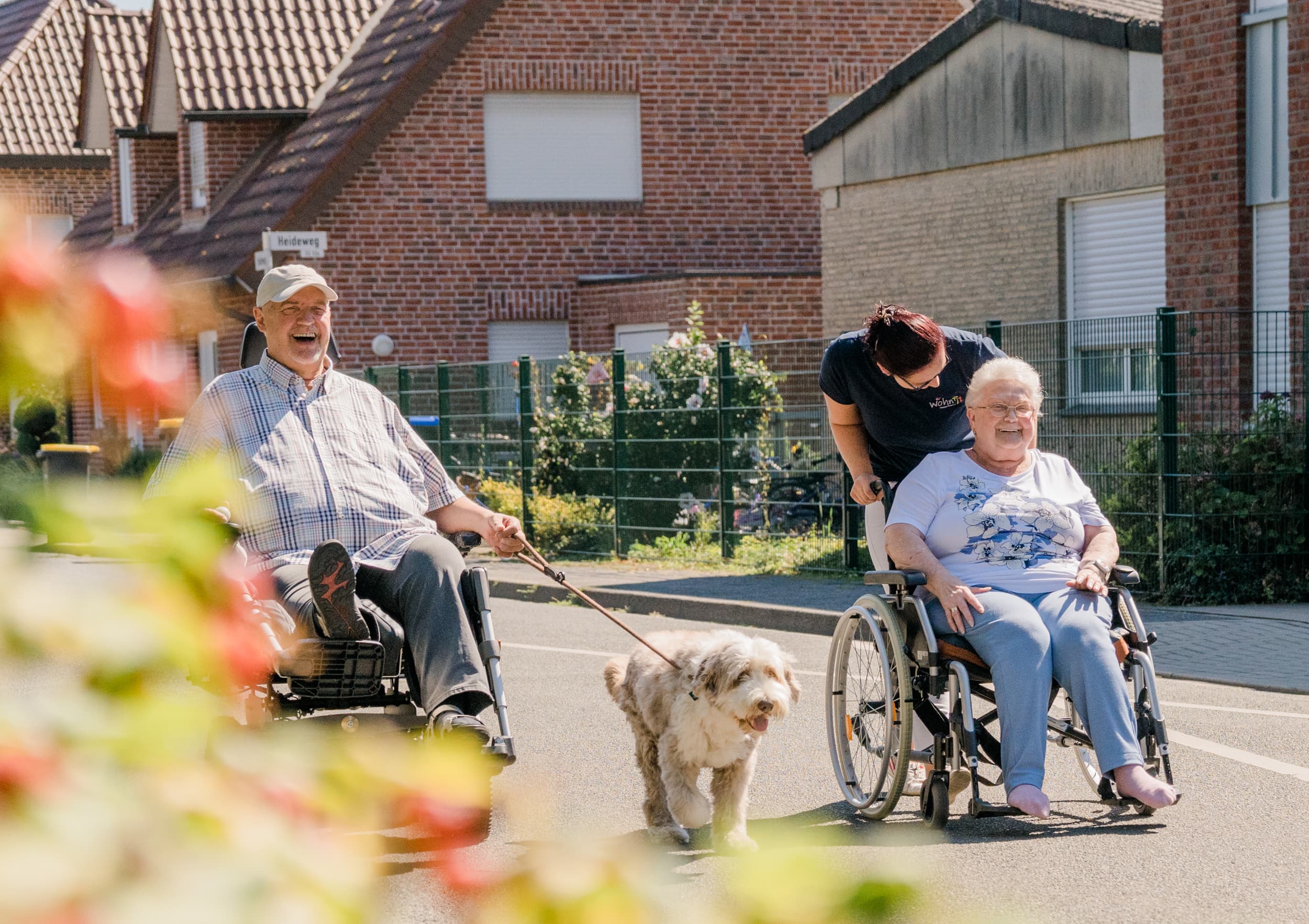 Pflegekraft schiebt Seniorin im Rollstuhl, daneben ein lachender Mann im E-Rollstuhl mit Hund.