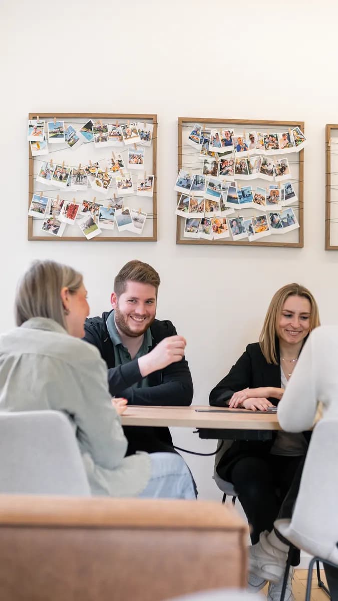 Drei Teammitglieder in einer Besprechung am Tisch, an der Wand Fotowände